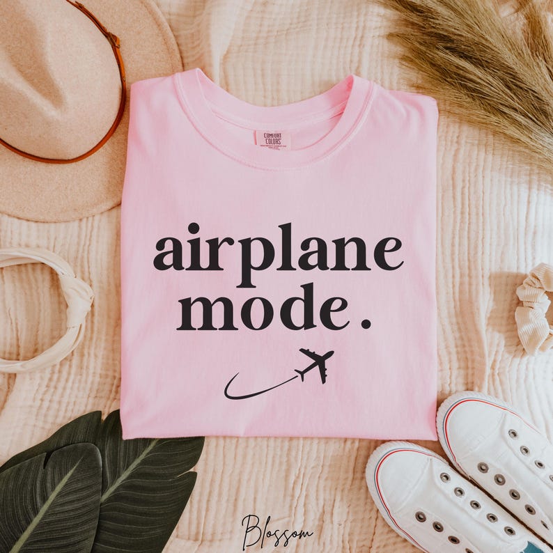 Op de afbeelding: Roze T-shirt met zwarte tekst die "vliegtuigmodus" zegt. Naast de tekst staat een klein vliegtuigpictogram.