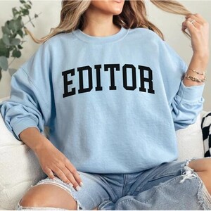 Pode incluir: Uma camisa de moletom azul claro com a palavra "EDITOR" impressa em letras pretas.