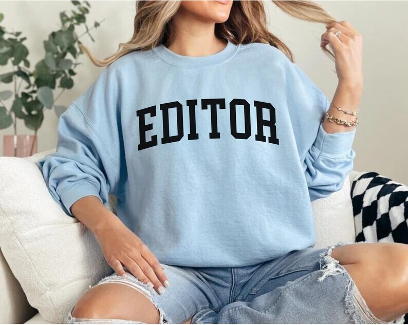 Pode incluir: Uma camisa de moletom azul claro com a palavra "EDITOR" impressa em letras pretas.