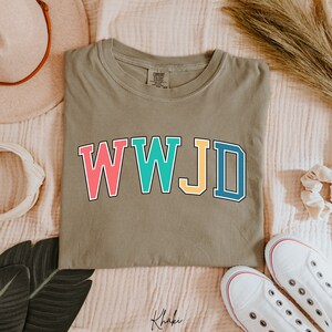 Op de afbeelding: Een kaki groen T-shirt met de tekst "WWJD" in kleurrijke blokletters. De letters zijn zwart omrand en elke letter is een andere kleur: roze, turquoise, geel en blauw.