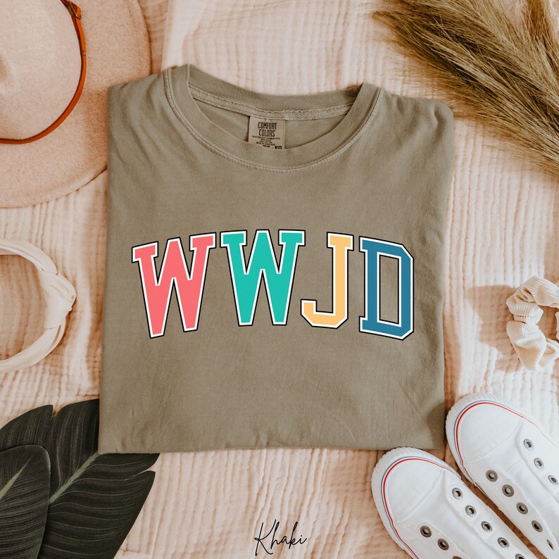 Op de afbeelding: Een kaki groen T-shirt met de tekst "WWJD" in kleurrijke blokletters. De letters zijn zwart omrand en elke letter is een andere kleur: roze, turquoise, geel en blauw.
