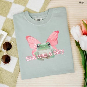 Op de afbeelding: Een lichtblauw t-shirt met een groene kikker die roze vlinder vleugels draagt. De tekst "She was a fairy" is op het shirt gedrukt.