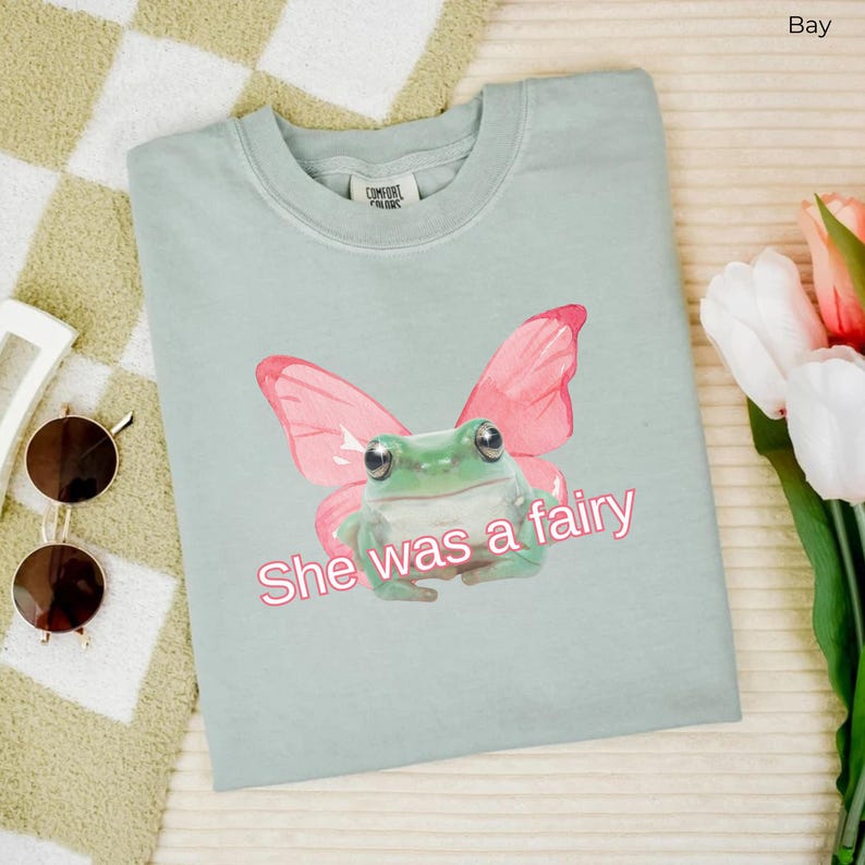 Op de afbeelding: Een lichtblauw t-shirt met een groene kikker die roze vlinder vleugels draagt. De tekst "She was a fairy" is op het shirt gedrukt.