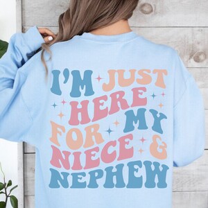 Pode incluir: Camisola azul clara com a frase "I'M JUST HERE FOR MY NIECE & NEPHEW" em letras de estilo retro. O texto &eacute; em tons de azul, rosa e laranja com detalhes de estrelas. Uma pe&ccedil;a de roupa casual e confort&aacute;vel.