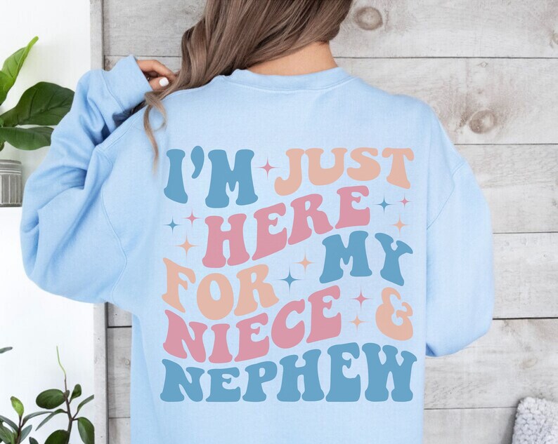 Pode incluir: Camisola azul clara com a frase "I'M JUST HERE FOR MY NIECE & NEPHEW" em letras de estilo retro. O texto &eacute; em tons de azul, rosa e laranja com detalhes de estrelas. Uma pe&ccedil;a de roupa casual e confort&aacute;vel.
