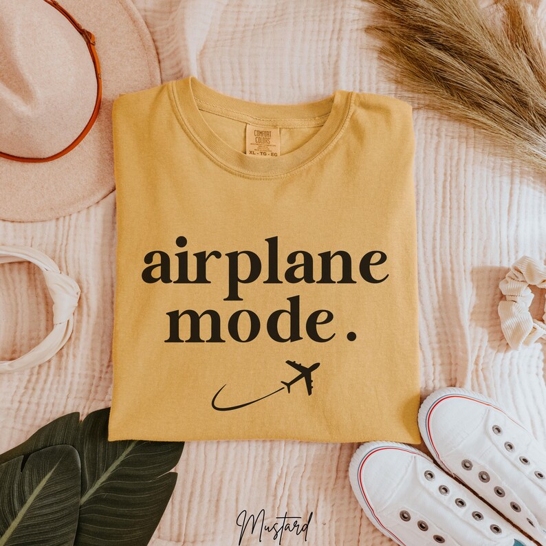Op de afbeelding: Een mosterdgeel T-shirt met de tekst "airplane mode." in zwarte letters. Een klein vliegtuigpictogram bevindt zich onder de tekst.