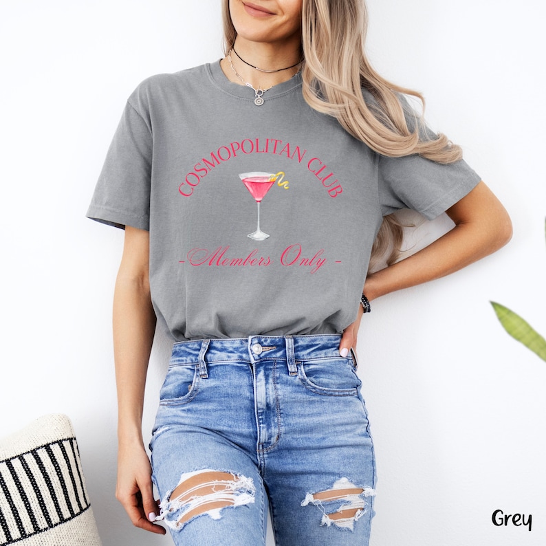 May include: A grey t-shirt with the text "Cosmopolitan Club - Members Only" in pink lettering. A pink cocktail glass with a lemon twist is pictured above the text.