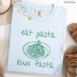 Puede incluir: Camiseta azul claro con un gr&aacute;fico verde de un plato de pasta y el texto "eat pasta run fasta".