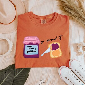 May include: A rust-colored t-shirt with a graphic of a jar of jam labeled "the gospel" and a slice of toast with jam spread on it. The text "go spread it." is printed above the toast.