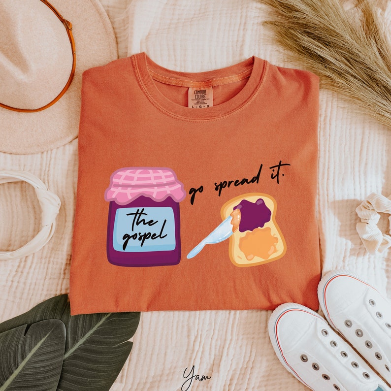 May include: A rust-colored t-shirt with a graphic of a jar of jam labeled "the gospel" and a slice of toast with jam spread on it. The text "go spread it." is printed above the toast.