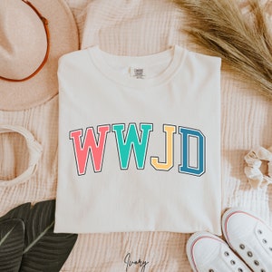 Op de afbeelding: Wit T-shirt met de tekst "WWJD" in kleurrijke blokletters. De letters zijn zwart omrand en elke letter heeft een andere kleur: roze, turquoise, geel en blauw.