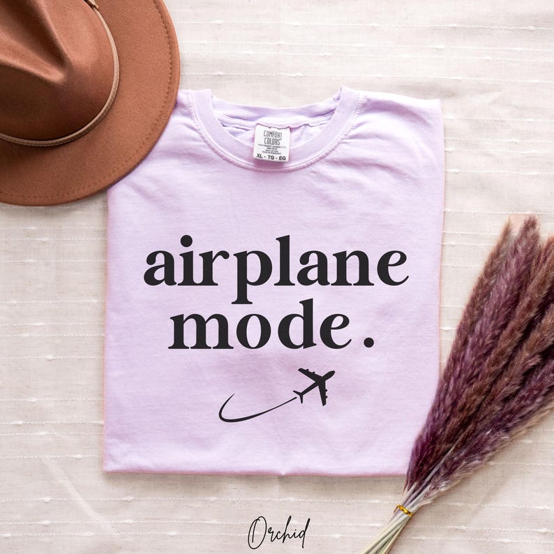 Op de afbeelding: Een lavendelkleurig T-shirt met de tekst "airplane mode." in zwarte letters. Een klein vliegtuigpictogram is onder de tekst gedrukt.