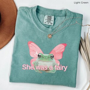 Op de afbeelding: Een lichtgroen T-shirt met een roze vlinder en een groene kikker. De kikker heeft grote ogen en draagt de vlindervleugels. De tekst "She was a fairy" is onder de kikker gedrukt.