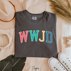 Op de afbeelding: Een donkergrijs T-shirt met de tekst "WWJD" in kleurrijke blokletters. De letters zijn zwart omrand en elke letter heeft een andere kleur: roze, turquoise, geel en blauw.