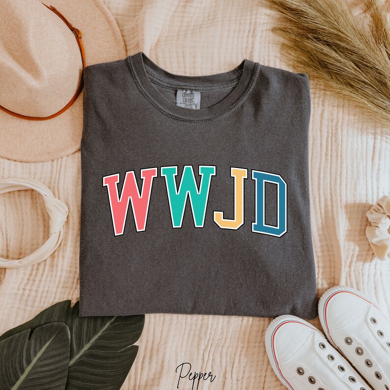 Op de afbeelding: Een donkergrijs T-shirt met de tekst "WWJD" in kleurrijke blokletters. De letters zijn zwart omrand en elke letter heeft een andere kleur: roze, turquoise, geel en blauw.