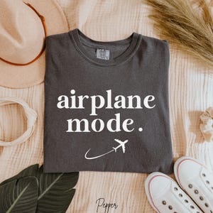 Op de afbeelding: Een donkergrijs T-shirt met de tekst "airplane mode." in witte letters. Een wit vliegtuigpictogram vliegt boven een witte gebogen lijn.