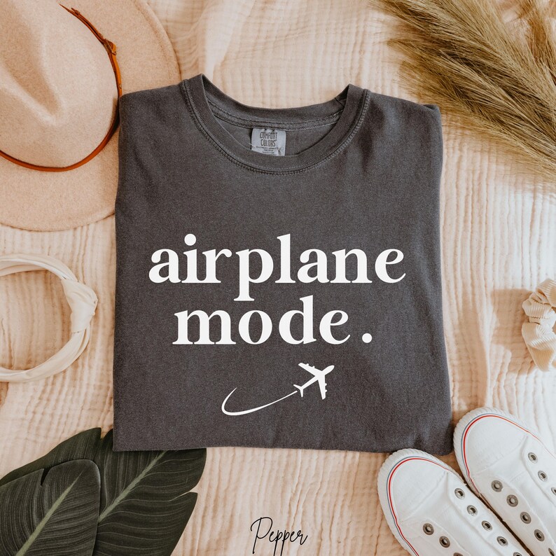 Op de afbeelding: Een donkergrijs T-shirt met de tekst "airplane mode." in witte letters. Een wit vliegtuigpictogram vliegt boven een witte gebogen lijn.