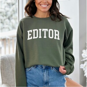 Pode incluir: Uma camisa de moletom verde com a palavra "EDITOR" impressa em branco na frente.