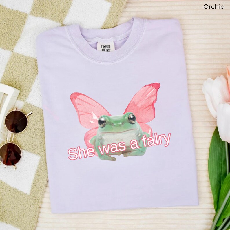 Op de afbeelding: Een lichtpaarse t-shirt met een groene kikker die roze vlinder vleugels draagt. De tekst "She was a fairy" is in roze onder de kikker gedrukt.