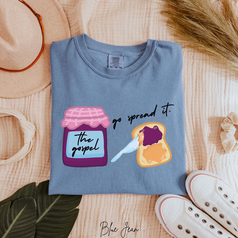 May include: A light blue t-shirt with a graphic of a jar of jam labeled "the gospel" and a slice of bread with jam spread on it. The text "go spread it." is printed on the shirt.