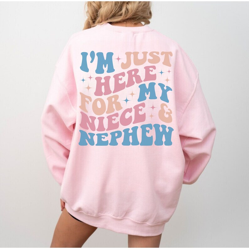 Pode incluir: Camisola rosa claro com o texto "I'M JUST HERE FOR MY NIECE & NEPHEW" em letras azuis, rosa e p&ecirc;ssego. O texto est&aacute; em uma fonte retr&ocirc; com detalhes de estrelas. Camisola de estilo casual.