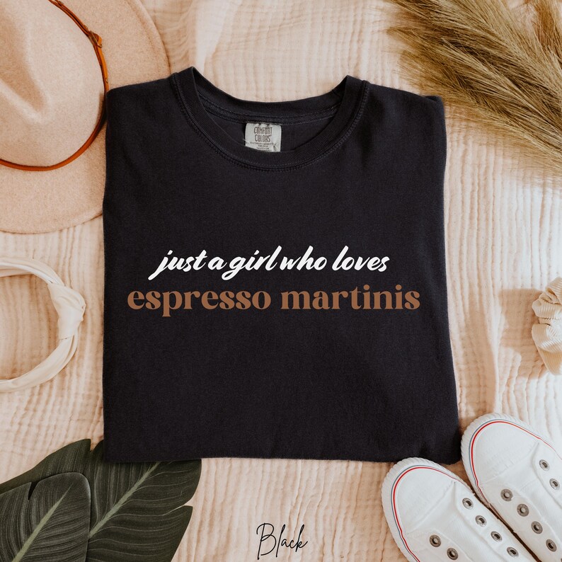 May include: Black t-shirt with the text "just a girl who loves espresso martinis" in white and brown lettering.