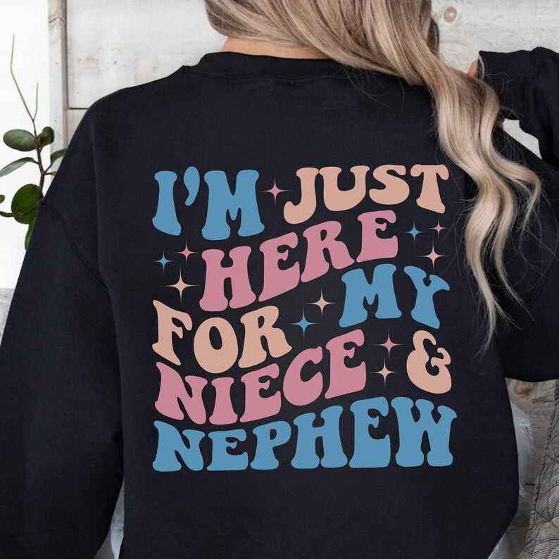 Pode incluir: Camisola preta com a frase "I'M JUST HERE FOR MY NIECE & NEPHEW" em uma fonte retr&ocirc;. O texto &eacute; em tons de azul, rosa e bege com detalhes de estrelas. Uma pe&ccedil;a de roupa casual e confort&aacute;vel.