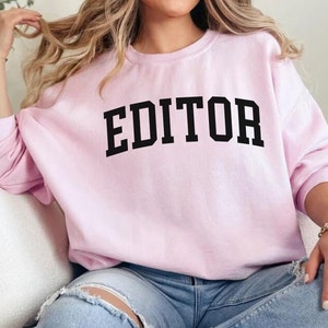 Pode incluir: Uma camisa de moletom rosa com a palavra "EDITOR" impressa em letras pretas.