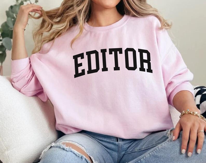 Pode incluir: Uma camisa de moletom rosa com a palavra "EDITOR" impressa em letras pretas.