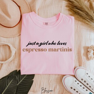 May include: Pink t-shirt with the text "just a girl who loves espresso martinis" in black and brown lettering.
