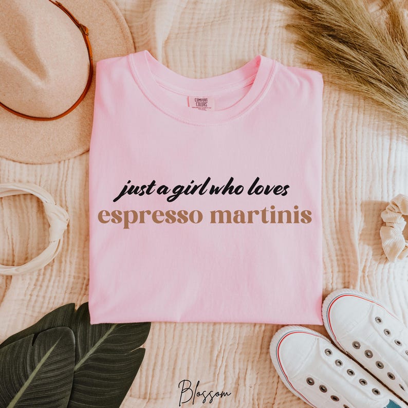 May include: Pink t-shirt with the text "just a girl who loves espresso martinis" in black and brown lettering.