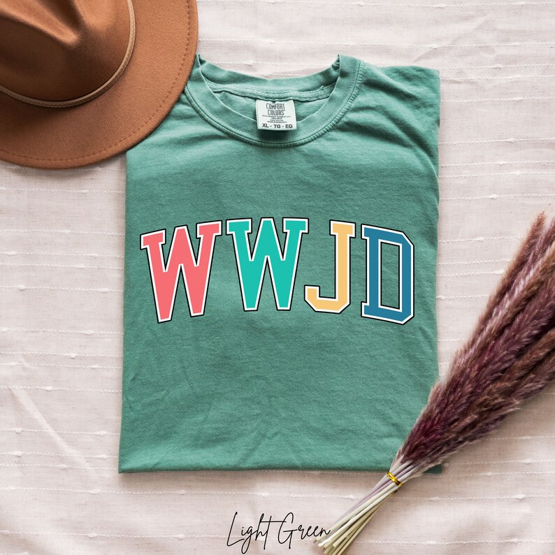 Op de afbeelding: Een lichtgroen T-shirt met de tekst "WWJD" in kleurrijke blokletters. De letters zijn zwart omrand en elke letter heeft een andere kleur: rood, turquoise, geel en blauw.