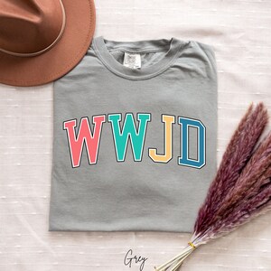 Op de afbeelding: Grijs T-shirt met de tekst "WWJD" in kleurrijke blokletters. De letters zijn zwart omrand.