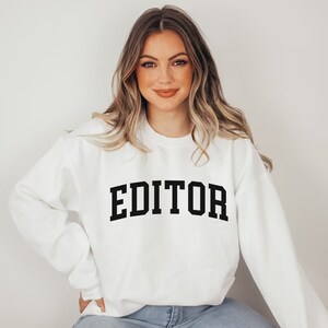 Pode incluir: Camisola branca com texto preto que diz "EDITOR" em uma fonte de estilo universit&aacute;rio.
