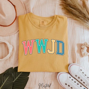 Op de afbeelding: Een mosterdgeel T-shirt met de tekst "WWJD" in kleurrijke blokletters. De letters zijn zwart omrand en elke letter heeft een andere kleur: roze, turquoise, geel en blauw.