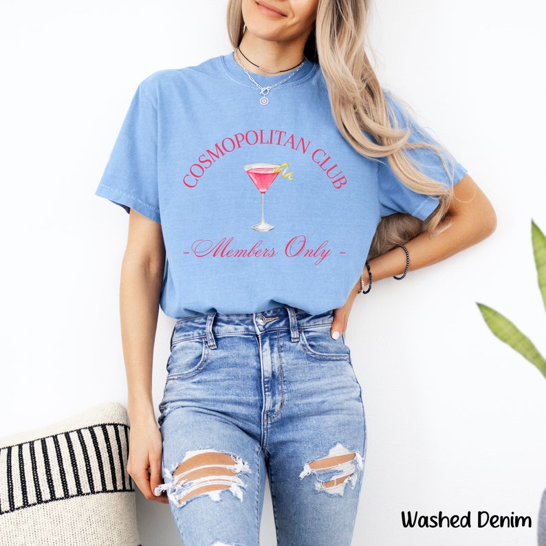 May include: A light blue t-shirt with the text "Cosmopolitan Club - Members Only" in pink lettering. A pink cocktail glass with a lemon twist is pictured above the text.