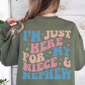 Pode incluir: Camisola verde-oliva com o texto "I'M JUST HERE FOR MY NIECE & NEPHEW" em letras azuis, rosa e p&ecirc;ssego. O texto est&aacute; em uma fonte retr&ocirc; e ondulada com estrelas.
