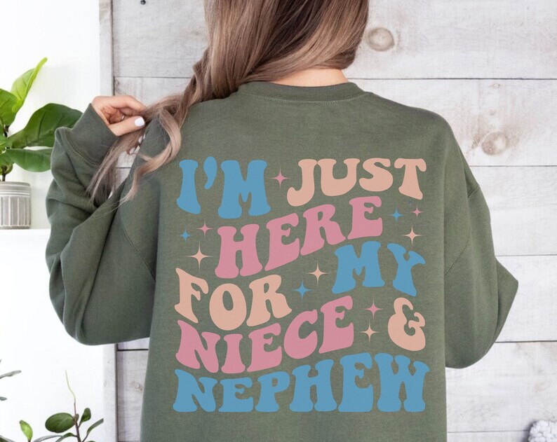 Pode incluir: Camisola verde-oliva com o texto "I'M JUST HERE FOR MY NIECE & NEPHEW" em letras azuis, rosa e p&ecirc;ssego. O texto est&aacute; em uma fonte retr&ocirc; e ondulada com estrelas.
