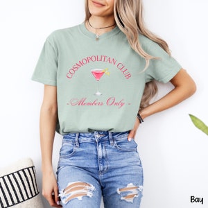 May include: A light green t-shirt with the text "COSMOPOLITAN CLUB" in pink and a pink cocktail glass with a lemon wedge. The text "Members Only" is written in cursive below the glass.