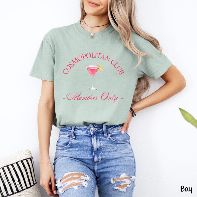 May include: A light green t-shirt with the text "COSMOPOLITAN CLUB" in pink and a pink cocktail glass with a lemon wedge. The text "Members Only" is written in cursive below the glass.