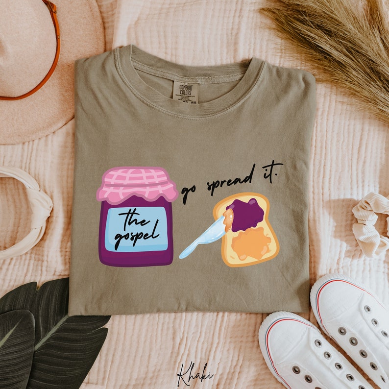 May include: Khaki t-shirt with a graphic of a jar of jam labeled "The Gospel" and a slice of toast with jam being spread on it. The text "go spread it." is above the toast.