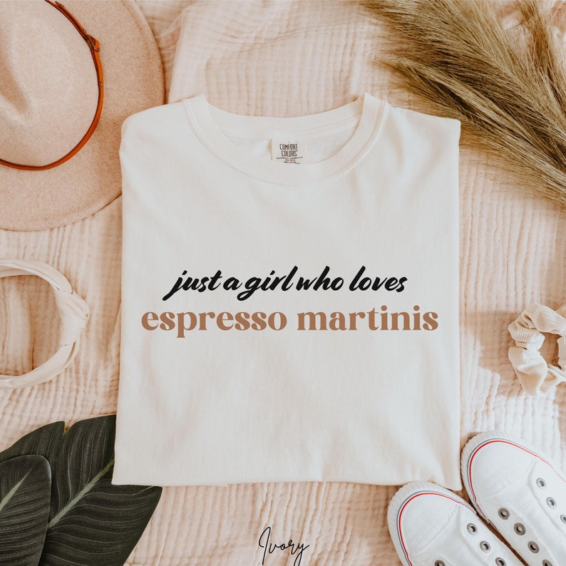 May include: White t-shirt with the text "just a girl who loves espresso martinis" in black and brown lettering.