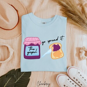 May include: Light blue t-shirt with a graphic of a jar of jam labeled "The Gospel" and a slice of toast with jam and peanut butter. The text "go spread it." is printed on the shirt.