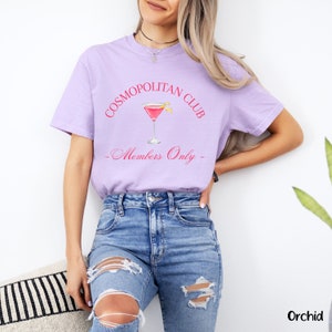 May include: A lavender t-shirt with the text "COSMOPOLITAN CLUB - Members Only -" in pink lettering. A pink cocktail glass with a lemon twist is pictured above the text.