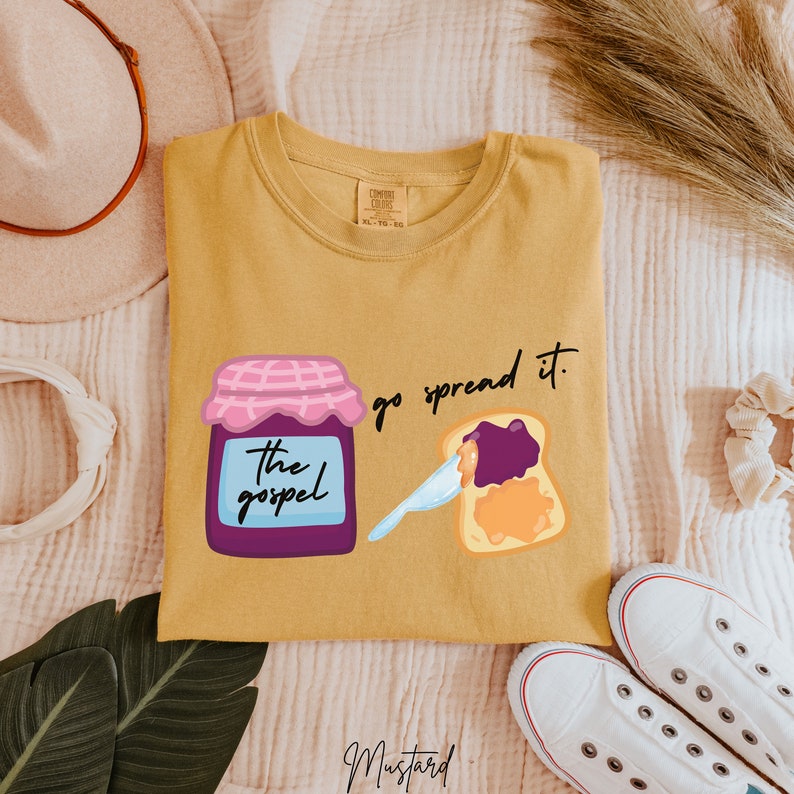 May include: A mustard yellow t-shirt with a graphic of a jar of jam labeled "The Gospel" and a slice of toast with jam spread on it. The text "go spread it." is printed on the shirt.