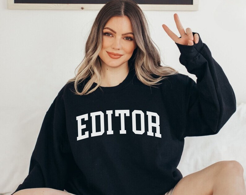 Pode incluir: Camisola de moletom preta com texto branco que diz "EDITOR" em uma fonte universit&aacute;ria.