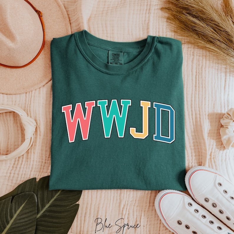 Op de afbeelding: Een donkergroen T-shirt met de tekst "WWJD" in kleurrijke blokletters. De letters zijn wit omrand. Het shirt ligt plat op een lichtroze, getextureerd oppervlak.