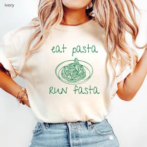 Puede incluir: Una camiseta blanca con un gr&aacute;fico verde de un plato de pasta y el texto "eat pasta run fasta".