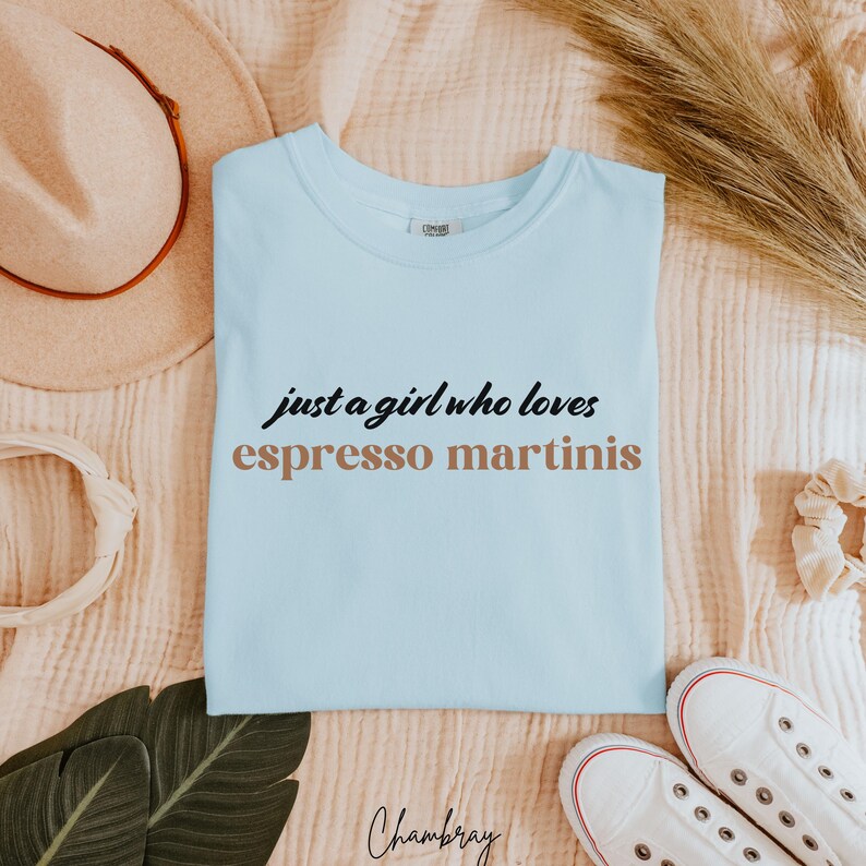May include: Light blue t-shirt with the text "just a girl who loves espresso martinis" in black and brown lettering.