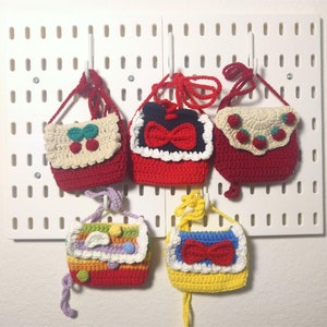 May include: Five small crocheted purses hanging on hooks. The purses are red, yellow, and cream with various colorful accents. Each purse has a different design, including cherries, bows, and rainbows.